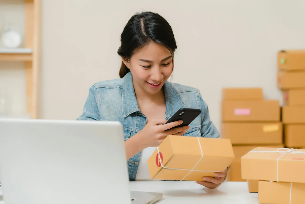Emprendedora sonriente preparando pedidos entre cajas, con el celular en la mano